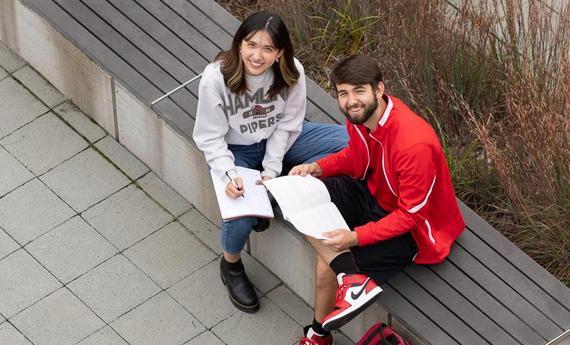 Two 侫Ƶ students sitting on a bench outside looking up at the camera smiling