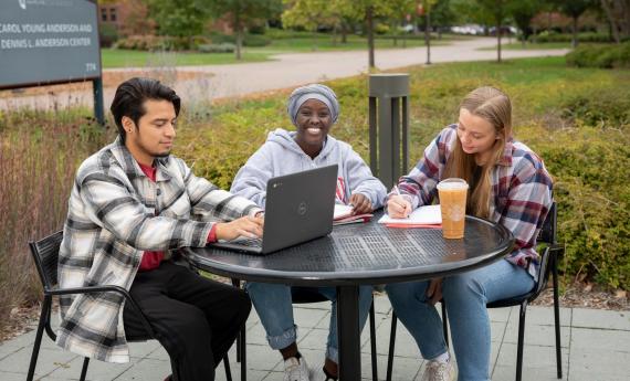 Three undergraduate students sitting outside at table on 侫Ƶ campus, one with laptop 