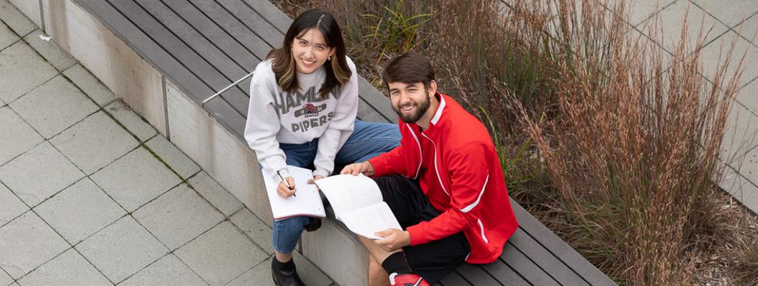 Two 侫Ƶ students sitting outside on campus with books and smiling up at the camera